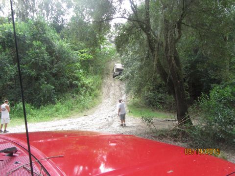 1995 Ford Bronco - A red vehicle is parked in the foreground, with a white SUV driving up a muddy hill in the background. Two men are standing on the side of…