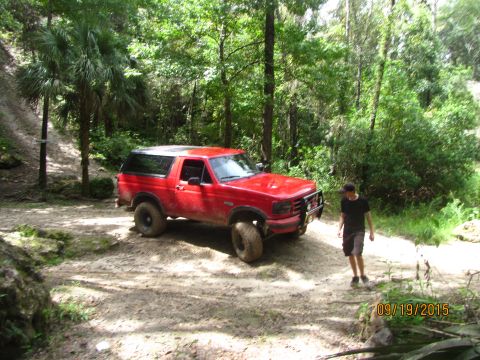 1995 Ford Bronco - A red Ford Bronco is parked on a dirt path in a wooded area. A person in a black t-shirt and shorts walks away from the vehicle.