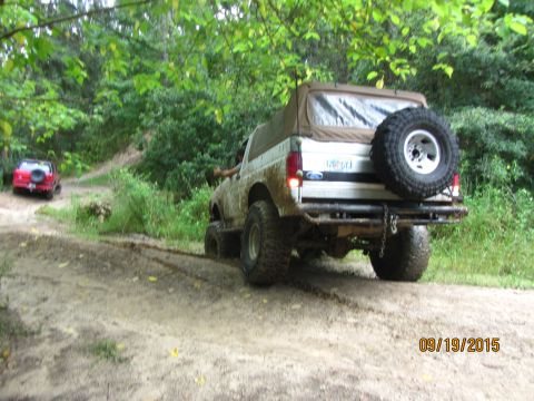1995 Ford Bronco - Two off-road vehicles are driving on a muddy trail through a wooded area. The foreground vehicle is a white Ford Bronco with a soft top and…