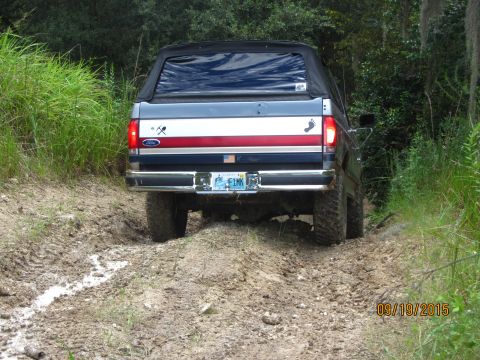 1995 Ford Bronco - The rear view of a dark blue Ford Bronco with a black soft top is shown driving on a muddy dirt road surrounded by greenery. The vehicle ha…