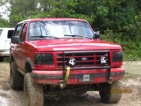 1995 Ford Bronco - A red 1995 Ford Bronco is parked off-road in a muddy area with trees in the background. The vehicle has large tires, aftermarket black head…