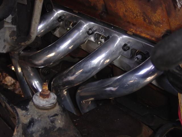 Chrome exhaust header tubes bolted to an engine block, viewed from under the wheel well area