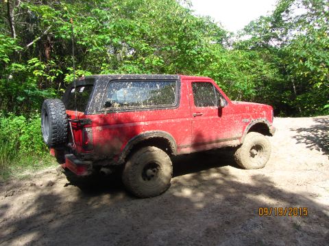 1995 Ford Bronco - A red, muddy 1995 Ford Bronco is parked on a dirt path surrounded by trees. The spare tire is mounted on the rear of the vehicle.