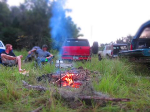 1995 Ford Bronco - Two men sit in camping chairs around a campfire with a grill over it. Several off-road vehicles are parked in the background.