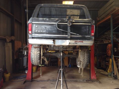 1979 Ford Bronco - The rear of a black Ford Bronco is raised on a red hydraulic lift in a workshop. The spare tire is hanging by straps below the vehicle.