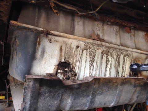 1979 Ford Bronco - This is a close-up, low-angle shot of the underside of a vehicle, focusing on a heavily rusted fuel tank and its mounting hardware. The tan…