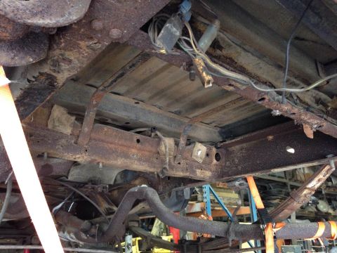 1979 Ford Bronco - The underside of a heavily rusted vehicle chassis is shown, with visible suspension components, exhaust pipes, and wiring. An orange strap…