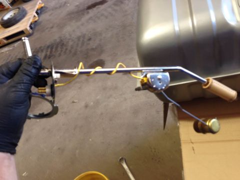 1979 Ford Bronco - A gloved hand holds a fuel sending unit with a yellow wire wrapped around it, positioned in front of a silver fuel tank. The sending unit h…