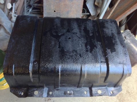 1979 Ford Bronco - A black, textured metal fuel tank is shown from a low angle, resting on a concrete surface. The tank has raised ridges and mounting holes a…