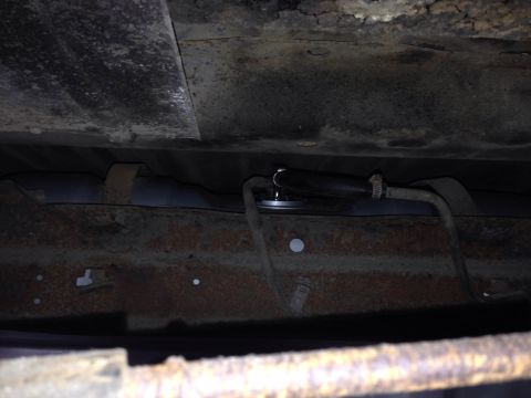 1979 Ford Bronco - The underside of a vehicle showing a fuel tank with a fuel line connected by a clamp. The frame and tank appear to be heavily rusted.