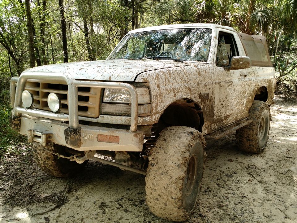 Mud-covered 1992 Ford Bronco with brush guard and large tires in a wooded area.