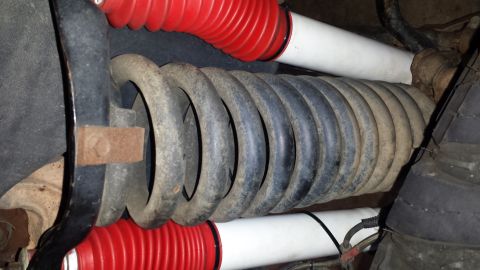 1996 Ford Bronco - Under-vehicle view of a front suspension showing a dirty gray coil spring seated in its perch alongside two white-bodied shock absorbers wi…
