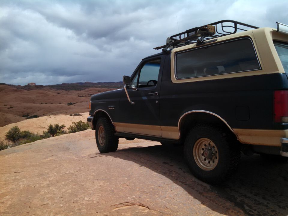 Dark green and tan Ford Bronco with roof rack parked on red slickrock under cloudy desert sky