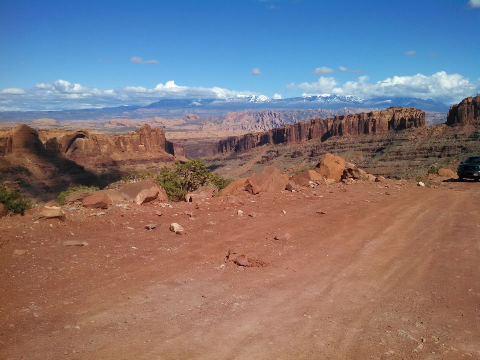 Desert landscape with red rock formations, distant snow-capped mountains, and a dark SUV parked on the right