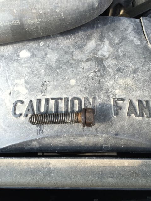 1989 Ford Bronco - A close-up view shows a bolt with a nut resting on a black plastic shroud with the embossed text "CAUTION FAN".