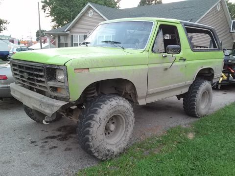 1986 Ford Bronco - A lifted, mud-covered 1980s Ford Bronco with oversized tires is parked on a gravel driveway in front of a house. The vehicle is bright gree…