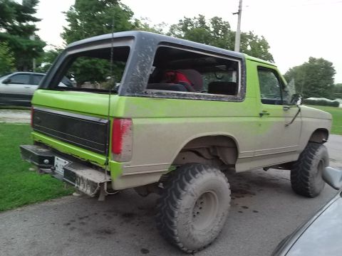 1986 Ford Bronco - The rear passenger side of a lifted, two-tone green and tan 1986 Ford Bronco is shown from a low angle. The vehicle has large off-road tire…