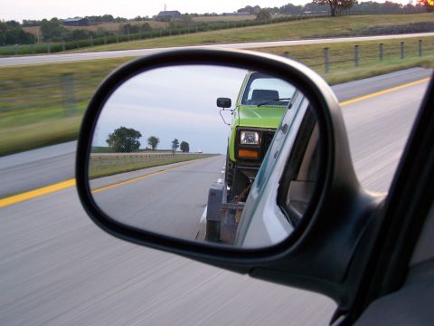 1986 Ford Bronco - A green pickup truck being towed on a trailer is reflected in a side-view mirror of a vehicle on a highway.