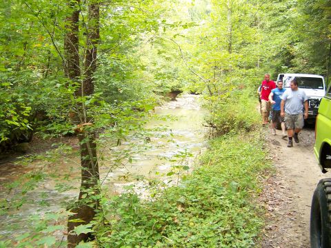 1986 Ford Bronco - A group of men walk along a dirt path next to a shallow, tree-lined creek. A white SUV is parked on the path, and the front of a green vehi…