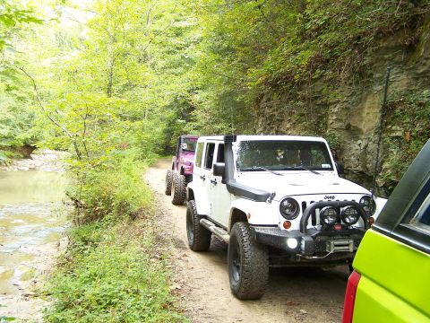 1986 Ford Bronco - A line of off-road vehicles, including a white Jeep Wrangler with a snorkel and winch, drives down a dirt path next to a creek. Another pur…