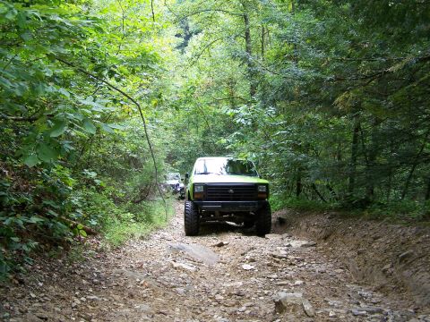 1986 Ford Bronco - A lifted, bright green Ford Bronco drives down a rocky, wooded trail. The vehicle is viewed from the front-right quarter angle.