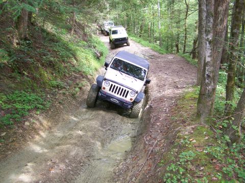 1986 Ford Bronco - A white Jeep Wrangler navigates a muddy, rutted trail in a forest setting. The vehicle is angled to the left, with its front right tire dee…