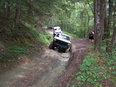 1986 Ford Bronco - A green 1986 Ford Bronco navigates a muddy trail through a forest, with a woman walking on the side of the path behind it.