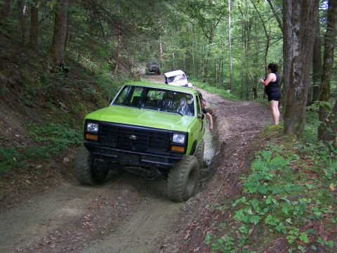 1986 Ford Bronco - A bright green Ford Bronco is driving through a muddy trail in a wooded area. A woman in a black top and shorts stands on the side of the t…