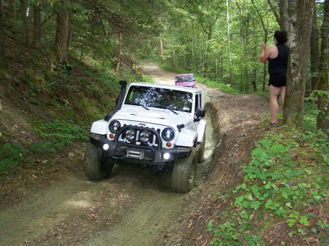 1986 Ford Bronco - A white Jeep Wrangler is driving through a muddy trail in a forest. A woman is standing on the side of the trail, looking at the Jeep.