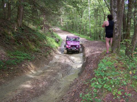 1986 Ford Bronco - A purple Jeep Wrangler is driving through a muddy water obstacle on a dirt trail in a forest. A woman stands on the side of the trail watch…