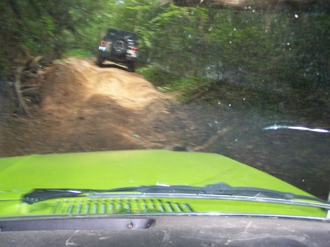 1986 Ford Bronco - The rear of a dark-colored SUV is visible driving up a dirt path in a wooded area. The front of a bright green vehicle is in the foreground…