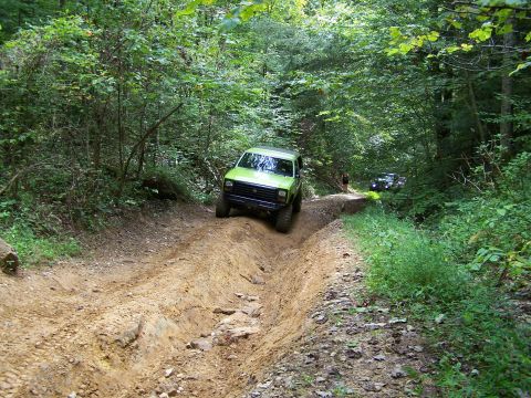 1986 Ford Bronco - A bright green 1986 Ford Bronco is driving off-road on a dirt path through a dense forest. Another vehicle is visible further down the trai…