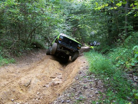 1986 Ford Bronco - A bright green 1986 Ford Bronco is navigating a steep, rutted dirt trail through a dense forest. The vehicle is tilted significantly to the…