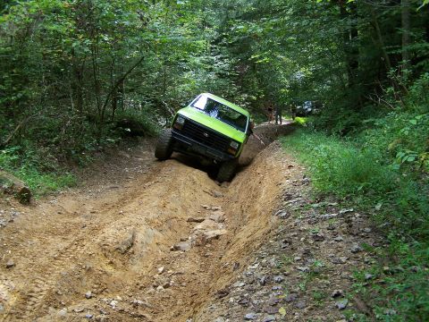 1986 Ford Bronco - A bright green 1986 Ford Bronco is navigating a steep, muddy off-road trail with dense green foliage on either side. The vehicle is angled…