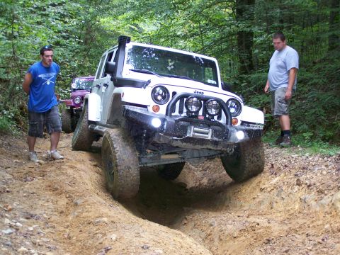 1986 Ford Bronco - Two Jeep Wranglers navigate a muddy, rutted trail in a wooded area. A man in a blue t-shirt stands to the left of the lead white Jeep, whil…