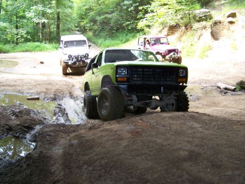 1986 Ford Bronco - A lifted green 1980s Ford Bronco is shown in a muddy off-road setting, with a white Jeep and a pink Jeep visible in the background.