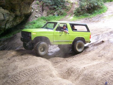 1986 Ford Bronco - A bright green 1980s Ford Bronco is navigating a muddy, off-road trail with a driver and passenger visible inside. The vehicle is angled up…