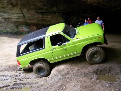 1986 Ford Bronco - A bright green Ford Bronco is stuck in mud inside a cave, with its tires sunk deep into the dirt. Several people are standing behind the ve…
