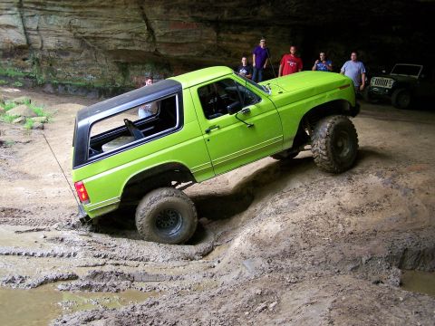 1986 Ford Bronco - A bright green Ford Bronco is navigating a muddy, uneven terrain inside a cave. Several people are gathered in the background, observing th…