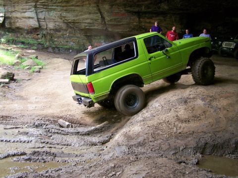 1986 Ford Bronco - A bright green 1980s Ford Bronco is navigating a muddy, off-road trail inside a cave or rock overhang. Several people are standing around w…