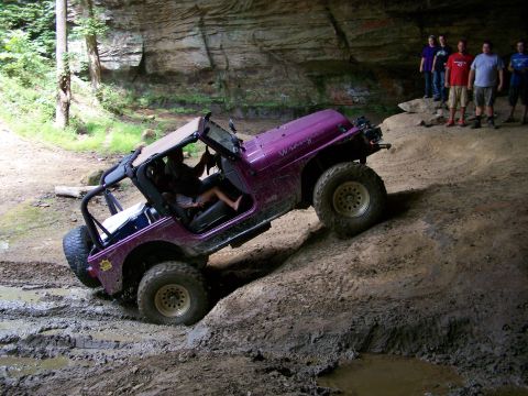 1986 Ford Bronco - A purple Jeep Wrangler is shown navigating a muddy, off-road trail inside a cave or rock overhang. Several people are standing in the backg…