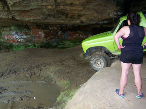 1986 Ford Bronco - A bright green Ford Bronco is parked inside a cave with graffiti on the walls, while a woman stands nearby looking at the vehicle.