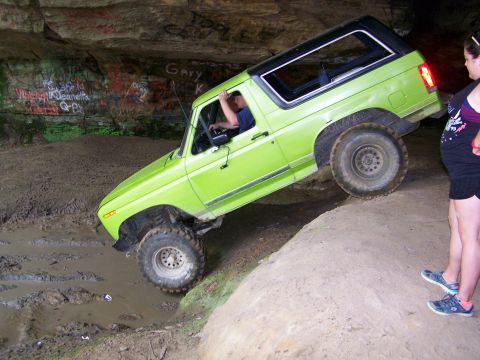 1986 Ford Bronco - A bright green 1986 Ford Bronco is navigating a muddy trail inside a cave with graffiti on the walls. The Bronco is tilted at a steep angle…