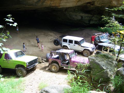 1986 Ford Bronco - A lime green Ford Bronco is parked on a dirt path next to a cave entrance, with several other Jeeps and people around.