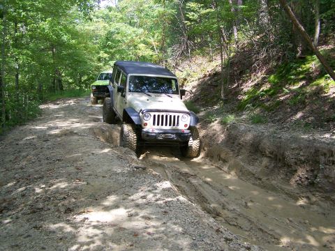 1986 Ford Bronco - Two off-road vehicles navigate a muddy rutted trail through a wooded area. The lead vehicle is a light-colored Jeep Wrangler, partially sub…
