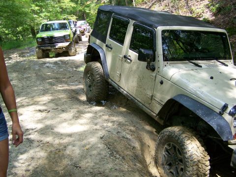 1986 Ford Bronco - A light-colored Jeep Wrangler is navigating a muddy, off-road trail, with a lime green Ford Bronco visible behind it. The Jeep is covered i…