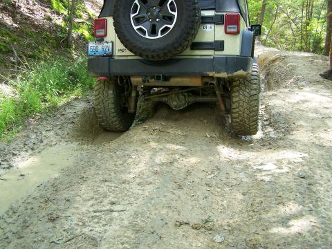 1986 Ford Bronco - The rear of a tan Jeep Wrangler is shown navigating a deep mud pit on a dirt trail. The vehicle is tilted to the left as its right rear tir…