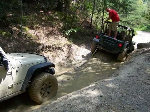 1986 Ford Bronco - Two Jeep Wranglers are stuck in a muddy ditch on a wooded trail. The front Jeep is tan and the rear Jeep is dark green, with a man standing…