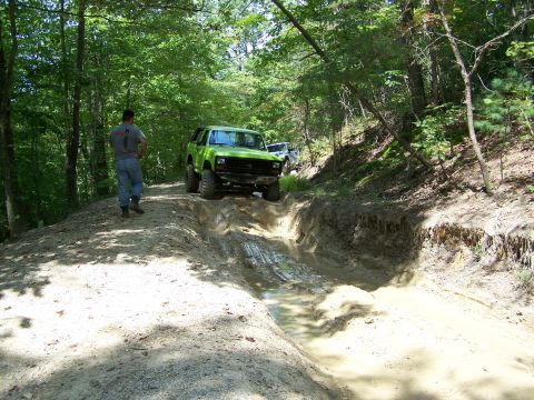 1986 Ford Bronco - A green Ford Bronco is navigating a deep mud pit on a wooded trail. A person in a grey t-shirt and jeans stands on the side of the trail wa…