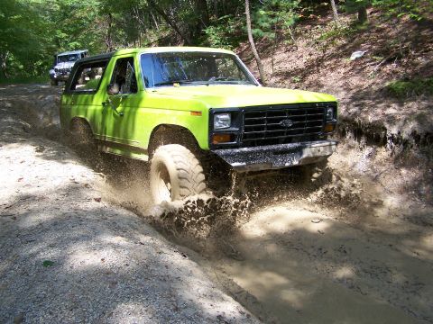 1986 Ford Bronco - A bright green 1986 Ford Bronco is driving through a muddy trail, kicking up a large splash of water and mud.